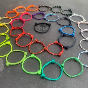 May include: A selection of brightly coloured rope bracelets, including orange, pink, green, and blue. Each bracelet has a knotted design and is displayed on a dark surface. The bracelets are a simple, minimalist style.