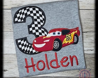 Op de afbeelding: Grijze t-shirt met een rode cartoon raceauto, nummer 95 en bliksemontwerp. Een groot nummer 3 met een geblokt vlagpatroon staat links. De naam "Holden" is rood geborduurd.
