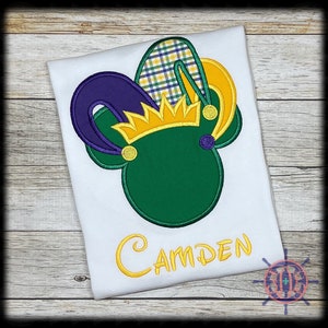 May include: White t-shirt featuring a green Mickey Mouse-shaped design with a gold crown and purple and yellow jester hat. The name "Camden" is embroidered in gold script below the design. The design is likely for a child.