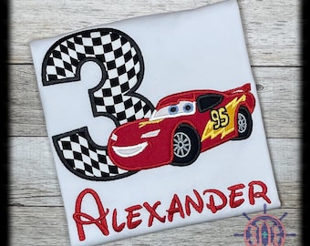 May include: White t-shirt with a red race car and the number 3 in a black and white checkered pattern. The name "Alexander" is embroidered in red script below the car. The car has the number 95 and a yellow lightning bolt.