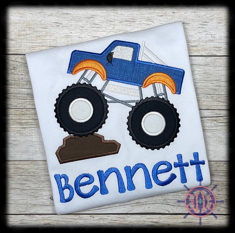 May include: White t-shirt featuring a blue monster truck design with orange accents and black tires. The name "Bennett" is embroidered in blue script below the truck. The design is centered on the shirt.