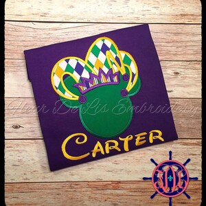 May include: Purple t-shirt with a green Mickey Mouse-shaped design wearing a Mardi Gras-style crown. The crown features yellow, white, green, and purple diamond patterns. The name "CARTER" is embroidered in yellow.