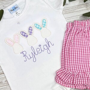 May include: White short-sleeved shirt with three embroidered bunny heads and the name "Ryleigh" in purple thread. Pink and white gingham ruffle shorts.