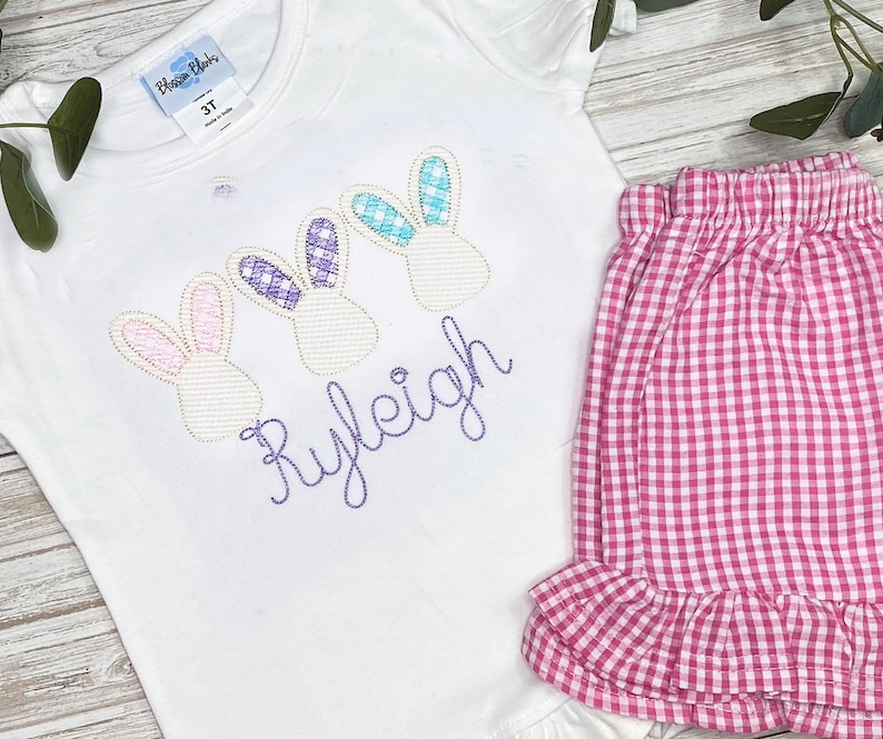 May include: White short-sleeved shirt with three embroidered bunny heads and the name "Ryleigh" in purple thread. Pink and white gingham ruffle shorts.