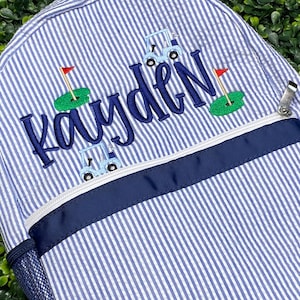 May include: A blue and white striped backpack with a navy blue zipper and trim. The backpack has a personalized embroidered name "Kayden" with golf cart and flag designs.