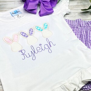 May include: A white t-shirt with a purple, pink, and blue bunny design and the name "Ryleigh" embroidered in purple. The shirt is paired with purple gingham shorts.