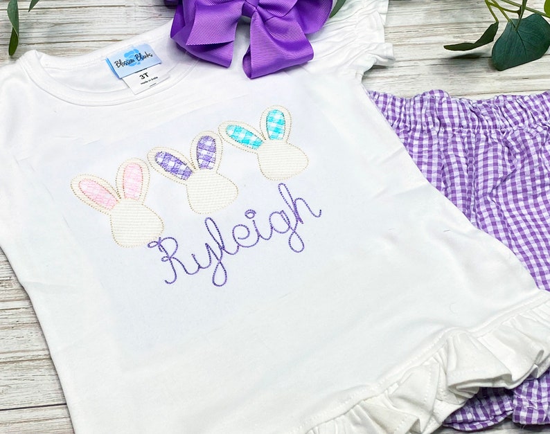 May include: A white t-shirt with a purple, pink, and blue bunny design and the name "Ryleigh" embroidered in purple. The shirt is paired with purple gingham shorts.