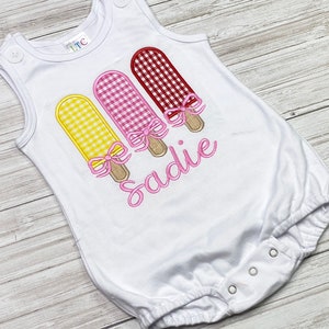 May include: White sleeveless onesie with three embroidered popsicles in yellow, pink, and red gingham patterns. The popsicles have pink bows and are arranged vertically. The onesie has the name "Sadie" embroidered in pink below the popsicles.