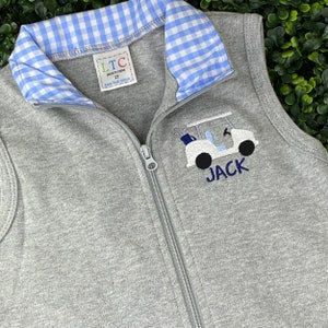 May include: Gray sleeveless zippered vest with a blue and white gingham collar. The vest has an embroidered golf cart with the name "JACK" underneath.