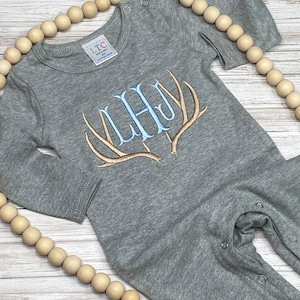 May include: Gray long-sleeve baby onesie with a monogram of the letters "L.H.J." embroidered in blue and white on a pair of brown antlers.