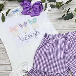 May include: A white short-sleeved shirt with embroidered purple bunnies and the name "Ryleigh" in purple. The shirt has a ruffled hem and is paired with purple gingham shorts.