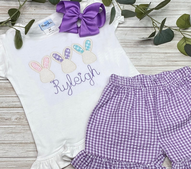 May include: A white short-sleeved shirt with embroidered purple bunnies and the name "Ryleigh" in purple. The shirt has a ruffled hem and is paired with purple gingham shorts.