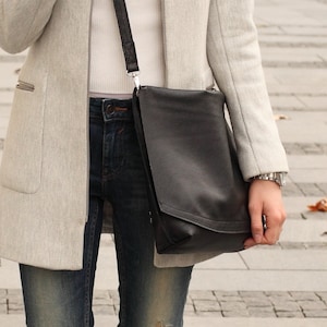 Może przedstawiać: Kobieta w szarym płaszczu i niebieskich dżinsach nosi czarną skórzaną torebkę na ramię.