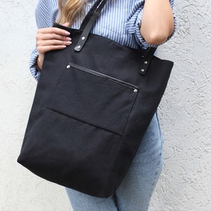 Op de afbeelding: Een zwarte canvas tote bag met een grote voorzak en zwarte leren handvatten. De tas heeft zilveren klinknagels op de handvatten en een ritszak aan de voorkant.