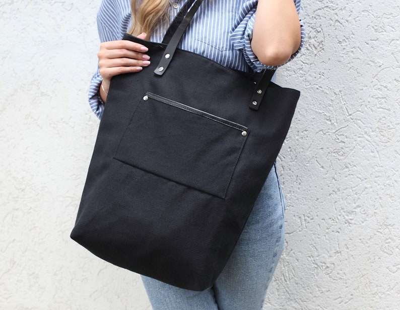 Op de afbeelding: Een zwarte canvas tote bag met een grote voorzak en zwarte leren handvatten. De tas heeft zilveren klinknagels op de handvatten en een ritszak aan de voorkant.