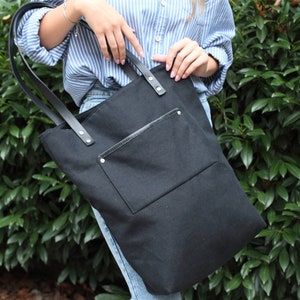 Op de afbeelding: Een zwarte canvas tote bag met een groot voorvak en leren banden. De tas heeft een ritssluiting en wordt door een persoon vastgehouden.