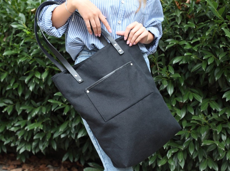 Op de afbeelding: Een zwarte canvas tote bag met een groot voorvak en leren banden. De tas heeft een ritssluiting en wordt door een persoon vastgehouden.