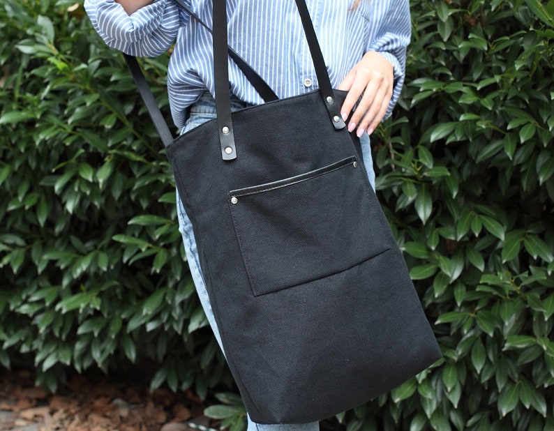 Op de afbeelding: Een zwarte canvas tote bag met een grote voorzak en zwarte leren banden. De tas heeft een ritssluiting en een klein lederen detail aan de voorkant.