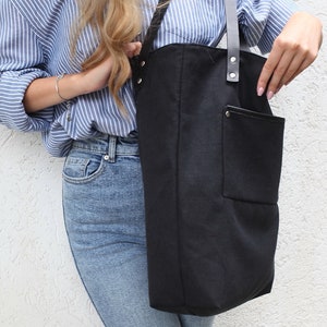 Op de afbeelding: Een zwarte canvas tote bag met een grote voorzak en zwarte leren banden. De tas wordt gedragen door iemand die een blauw-wit gestreept shirt en een blauwe jeans draagt.