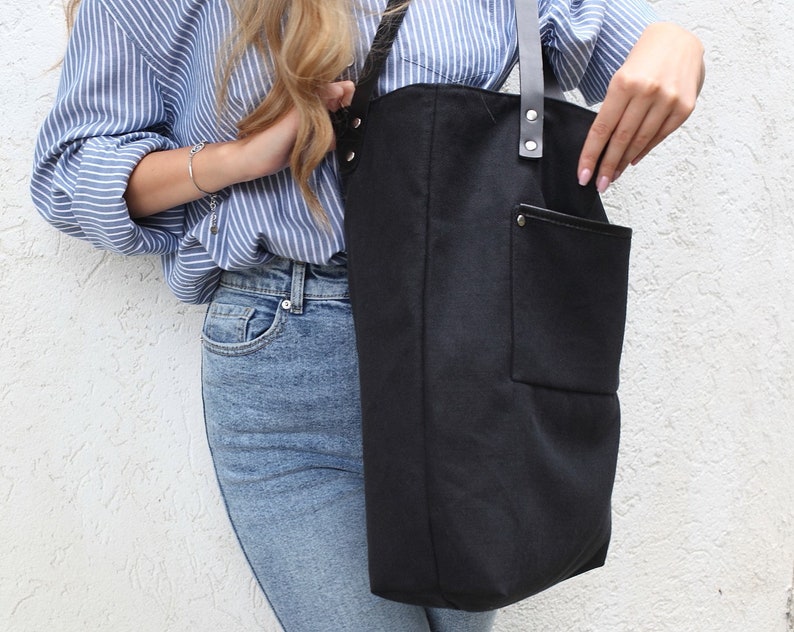Op de afbeelding: Een zwarte canvas tote bag met een grote voorzak en zwarte leren banden. De tas wordt gedragen door iemand die een blauw-wit gestreept shirt en een blauwe jeans draagt.