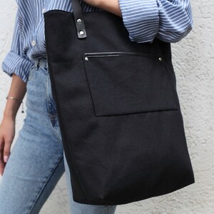 Op de afbeelding: Een zwarte canvas tote bag met een grote voorzak en een leren handvat. De tas wordt gedragen door iemand die een blauw-wit gestreept shirt en een blauwe jeans draagt.