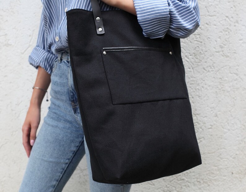 Op de afbeelding: Een zwarte canvas tote bag met een grote voorzak en een leren handvat. De tas wordt gedragen door iemand die een blauw-wit gestreept shirt en een blauwe jeans draagt.