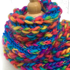 Può includere: Una vivace sciarpa a infinito lavorata a mano in un arcobaleno di colori, tra cui blu, giallo, arancione e viola. Il design a maglia grossa è esposto su una testa di manichino in legno, evidenziando la consistenza e lo stile della sciarpa. Un accessorio perfetto per aggiungere colore.