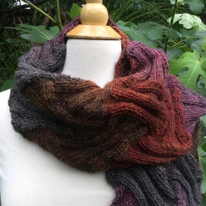 May include: A chunky knit infinity scarf featuring a variegated color palette of deep charcoal, burnt orange, and rich plum. The scarf displays a prominent cable knit texture throughout, creating a thick, layered appearance around the neck.