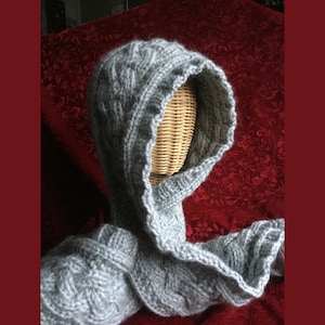 Könnte beinhalten: Ein grauer gestrickter Kapuzen-Schal mit einem Zopfmuster. Der Schal ist über einen Mannequin-Kopf gelegt.