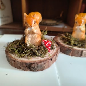 May include: Two small orange squirrel figurines standing on circular wood slices. Each squirrel is surrounded by green moss and a tiny red mushroom with white spots. The wood slices show natural bark edges and visible tree ring patterns.