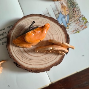 May include: An orange mushroom-shaped hair clip with a metal alligator clasp attached to the top, resting alongside a separate, smaller, light orange mushroom-shaped piece on a circular wooden slice with visible tree rings and bark edges.