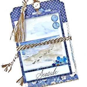 May include: A blue and white scrapbook page with a watercolor painting of a beach scene. The page is decorated with a brown twine bow, a blue button, and blue and white beads. The text "Seaside" is written in black cursive on the page. The page is from Button Farm.