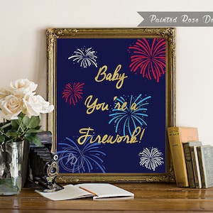 May include: A blue and gold sign with the text "Baby you're a firework!" surrounded by red, white, and blue fireworks. The sign is in a gold frame and is sitting on a wooden table.