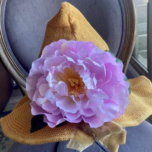 May include: A mustard yellow knitted sun hat adorned with a large, light purple peony flower. The flower has a yellow center and green leaves. A gold velvet ribbon is tied around the hat.