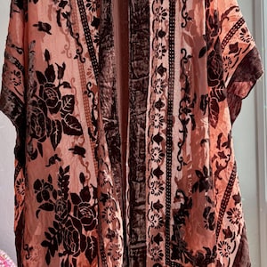 May include: A peach-colored kimono-style robe with a floral pattern in dark brown. The robe has a velvet texture and a fringe trim at the bottom. The design includes vertical stripes and floral motifs.
