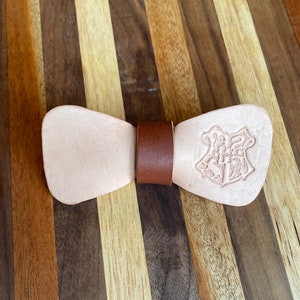 May include: A light brown leather bow tie with a brown leather strap and a Hogwarts crest embossed on one side.
