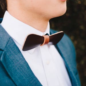 May include: A man wearing a blue suit jacket and a white shirt with a brown leather bow tie.