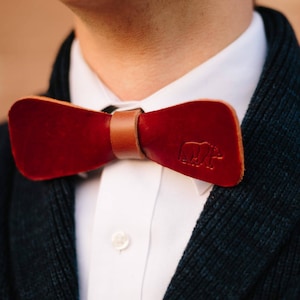 May include: A burgundy leather bow tie with a brown leather strap. The bow tie has a bear logo embossed on the right side. The bow tie is worn over a white collared shirt and a dark blue sweater.