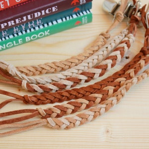 May include: Five braided leather straps with metal clips. The straps are brown and beige and are braided together in a pattern. The straps are lying on a wooden surface.