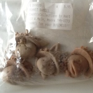 May include: Four unfinished, unpainted, beige ceramic doll heads with bonnets. The doll heads are packaged in a clear plastic bag with a price tag that reads "S 0H9005 BONNET BABIES 5.95 CountryCollections by Hart 13 M.Main, Moore 799-0773 THANKS for your BUSINESS!!"