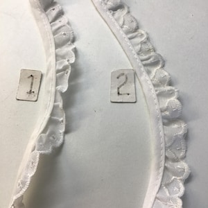 May include: Two pieces of white lace trim with scalloped edges and decorative cutouts. The trim is on a white background, with small tags labeled "1" and "2" visible.