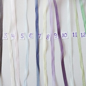May include: Twelve different colored ribbon samples, numbered 1 through 12. The ribbons are arranged horizontally, with the first ribbon being gray, the second white, the third pink, the fourth light blue, the fifth dark blue, the sixth cream, the seventh lavender, the eighth purple, the ninth white, the tenth light green, the eleventh yellow, and the twelfth light green.