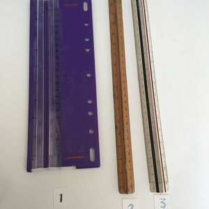 May include: Three rulers, one purple with the word "FISKARS" printed on it, one wooden, and one white with black and red markings. The rulers are numbered 1, 2, and 3.