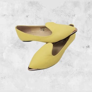May include: A pair of pale yellow slip-on flats. The shoes have a pointed toe with a gold-colored metal accent and a low heel. The interior of the shoe is a light beige color.