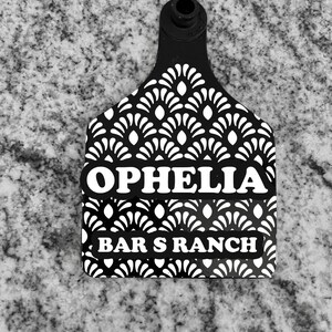 May include: Black and white plastic cattle ear tag with a white geometric pattern. The tag has the text "OPHELIA" and "BAR S RANCH" printed on it.
