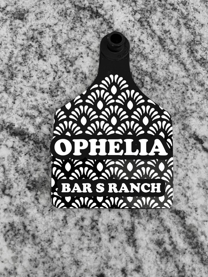May include: Black and white plastic cattle ear tag with a white geometric pattern. The tag has the text "OPHELIA" and "BAR S RANCH" printed on it.