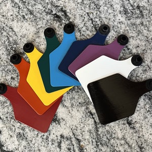 May include: A set of nine colourful plastic ear tags with black rubber rings. The tags are arranged in a fan shape, with the darkest tag at the bottom and the lightest tag at the top.