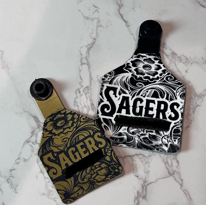 Puede incluir: Dos etiquetas para orejas de ganado de color negro y dorado con un dise&ntilde;o floral y la palabra "Sagers" impresa en ellas.