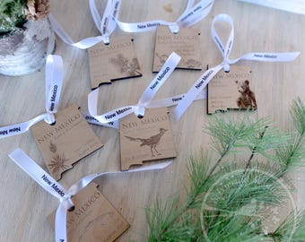 May include: Six wooden ornaments shaped like the state of New Mexico, each with a different design. The ornaments are decorated with images of a roadrunner, a pinon tree, and other state symbols. Each ornament has a white ribbon attached.