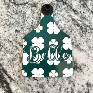 May include: Green plastic livestock ear tag with a white four-leaf clover pattern and the name "Belle" in white cursive lettering.